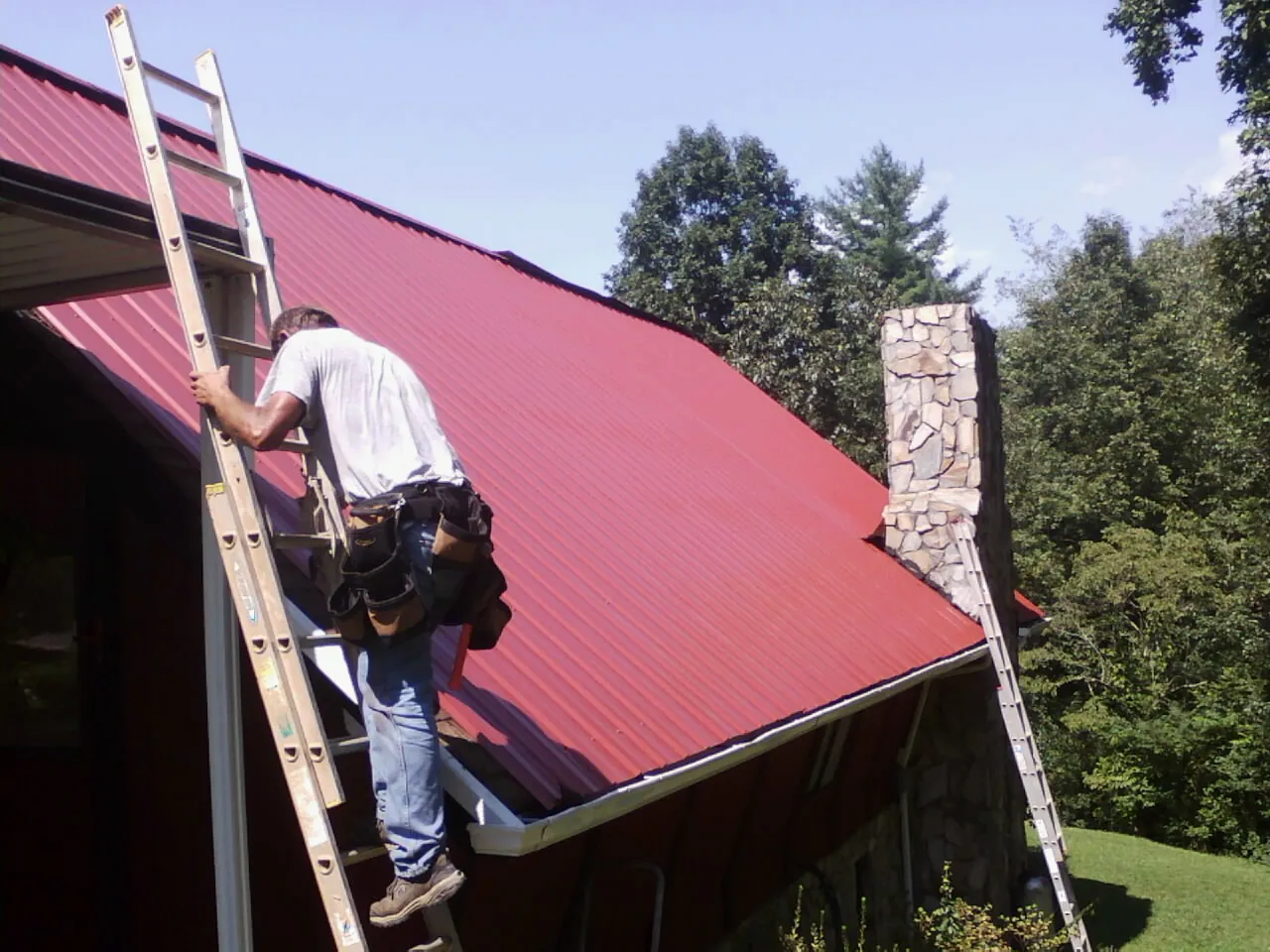 Roofer on a ladder beside a red metal roof and stone chimney — Soffit & Fascia Repair by Roofing Brothers in Pocasset