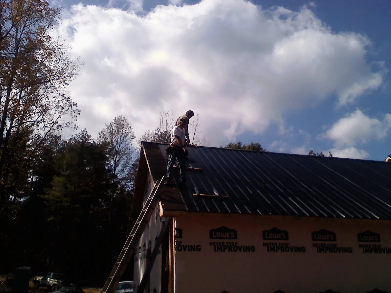 Roofer fastening dark metal panels high against a cloudy sky, from a Roof Inspection job in Pocasset